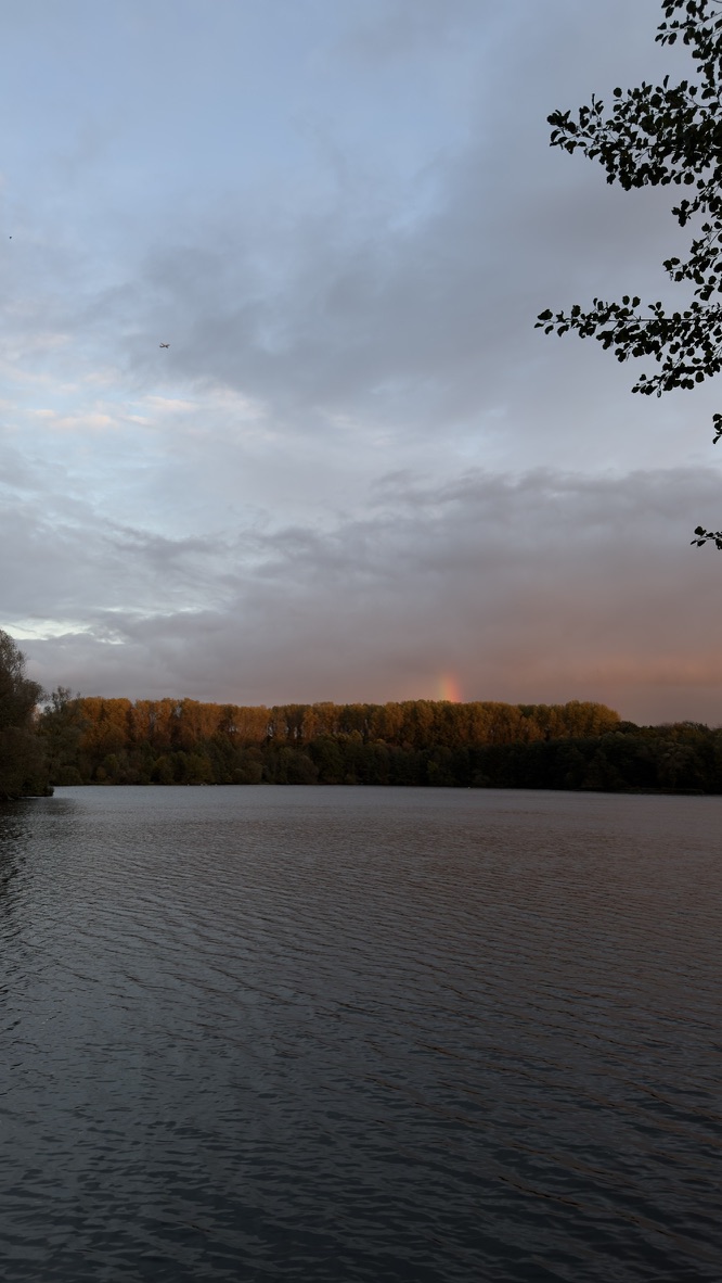 Schwacher Regenbogen über dem Erlensee in der Abenddämmerung